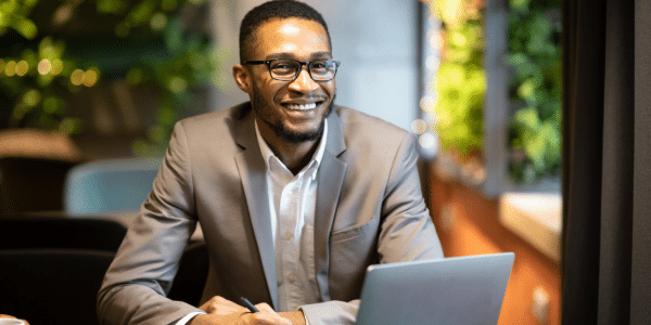 Smiling man in glasses and suit working on a laptop in an office with greenery in the background. The setting is professional and welcoming.